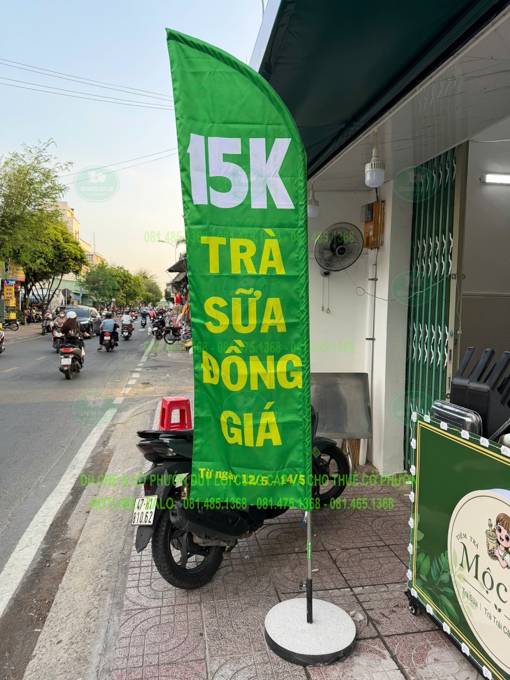 Cờ cánh buồm màu xanh có nội dung trà sữa đồng giá 15K, dựng bằng đế tròn trước cửa hàng.