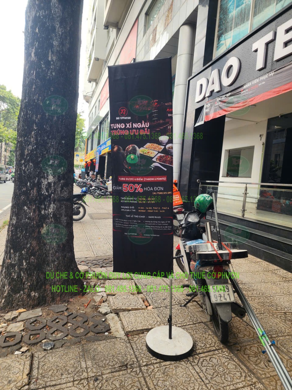 Cờ chữ nhật màu đen đặt gần cây và mặt tiền nhà hàng, bên cạnh khu vực xe đỗ.