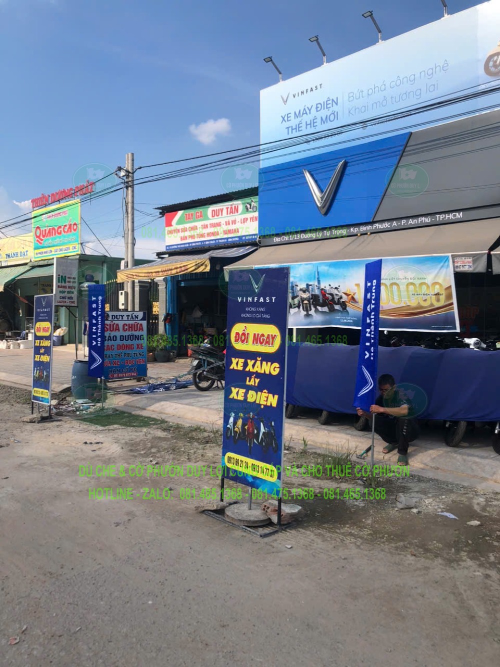 Các cờ chữ nhật nền xanh và xanh–vàng mang nội dung quảng cáo trước cửa hàng xe VinFast.