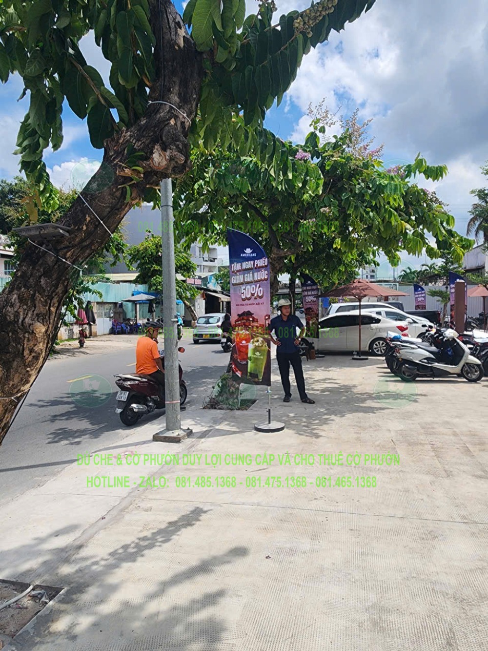 Một bộ cờ dựng dưới tán cây ở hướng tiếp cận, phía sau là khu vực xe và cửa hàng.