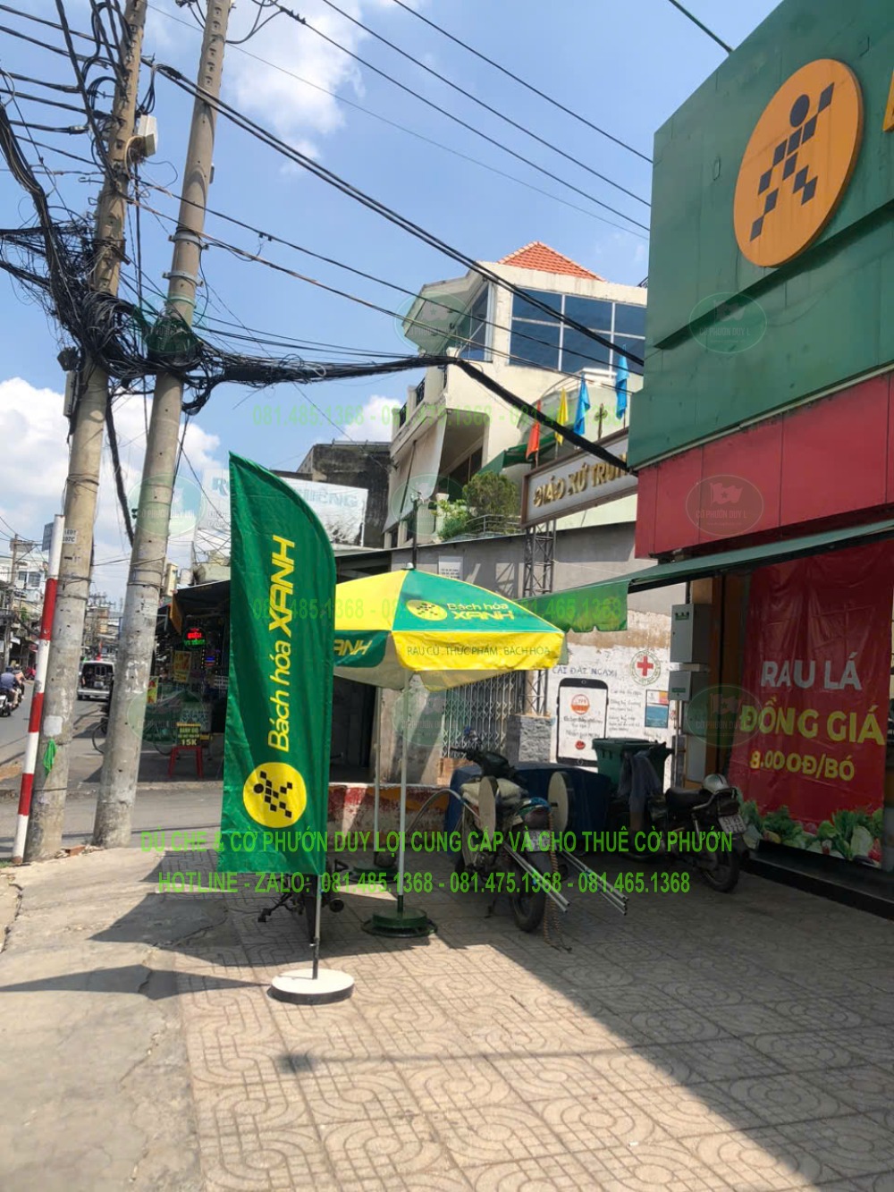 Cờ cánh buồm Bách Hóa Xanh nền xanh, chữ và biểu trưng vàng, đặt cùng bảng hiệu và dù che đồng bộ nhận diện.