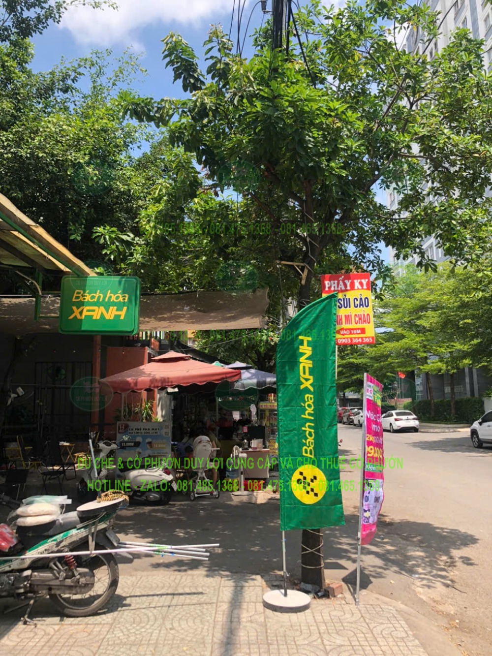 Cờ nền xanh dựng dưới tán cây, phía sau là biển hiệu Bách Hóa Xanh và khu vực điểm bán.