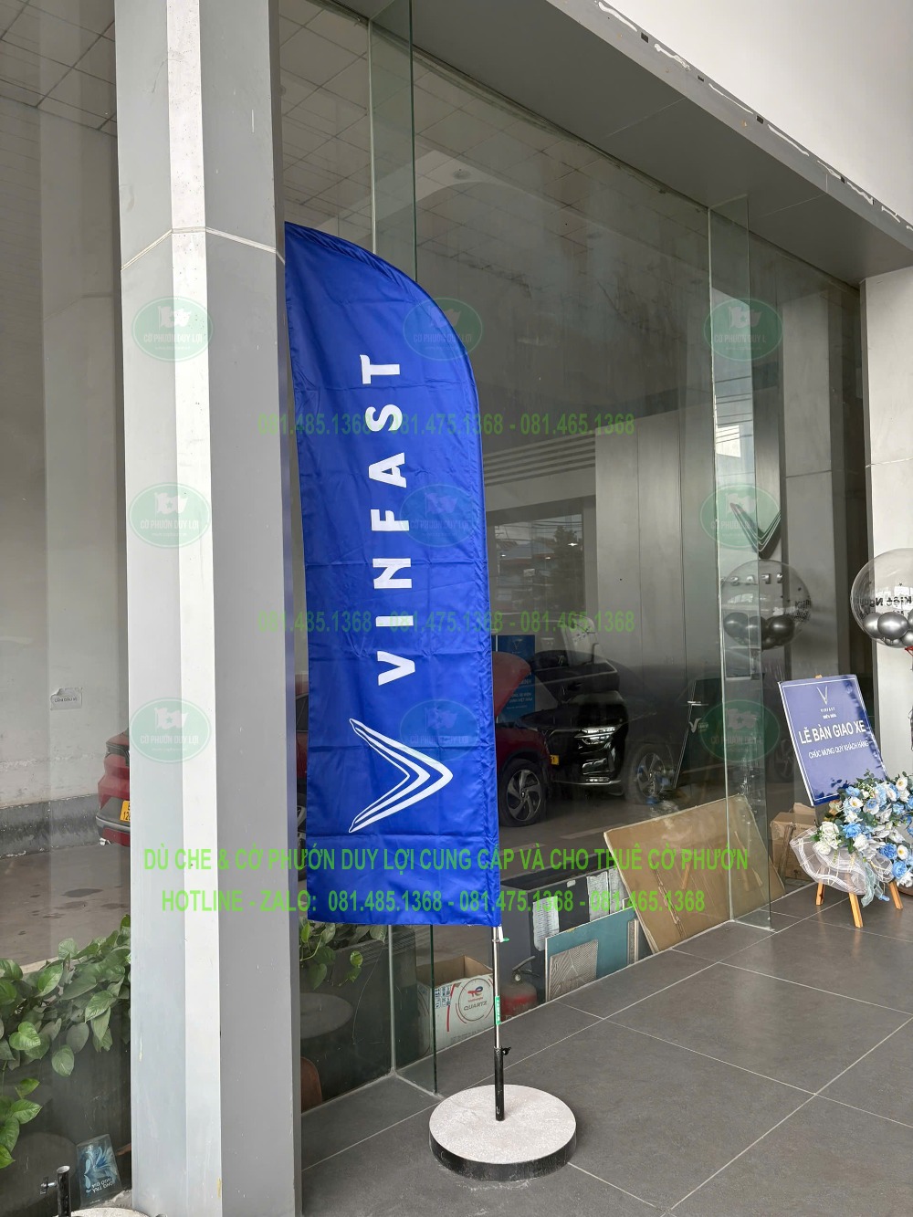 Cờ cánh buồm nền xanh mang tên và biểu trưng VinFast, dựng bằng đế tròn cạnh mặt kính showroom.