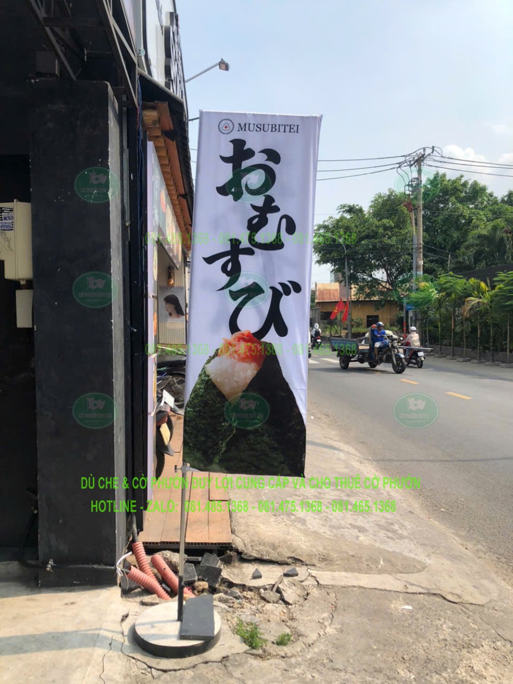 Cờ chữ nhật nền trắng in chữ Nhật và hình sản phẩm, đặt trước cửa hàng sát hướng người đi đường tiếp cận.