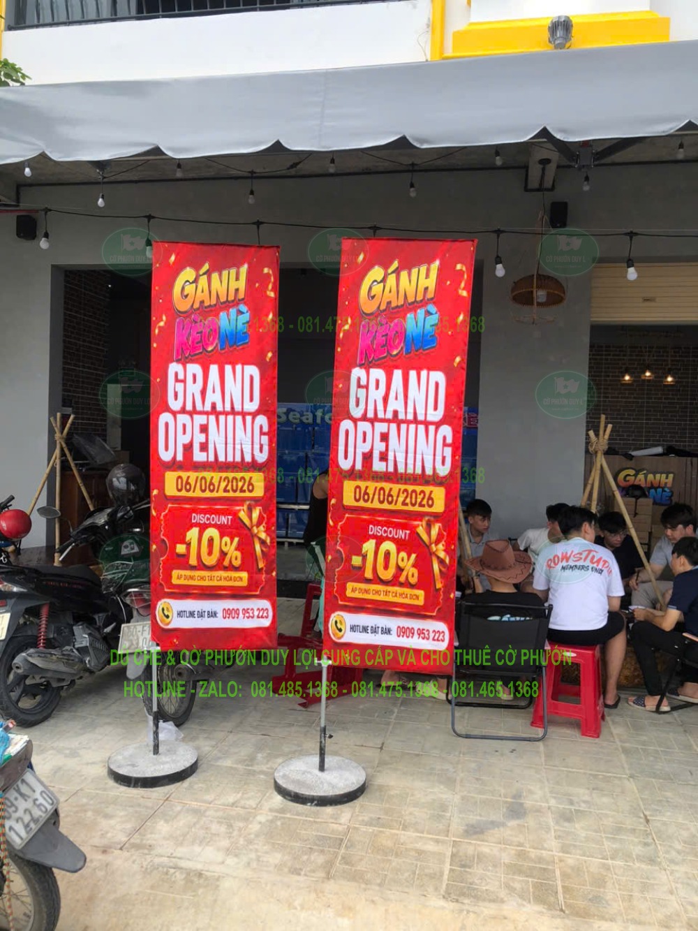 Hai cờ chữ nhật màu đỏ mang nội dung khai trương đặt trước quán ăn; chương trình trên lá thuộc thời điểm chụp.
