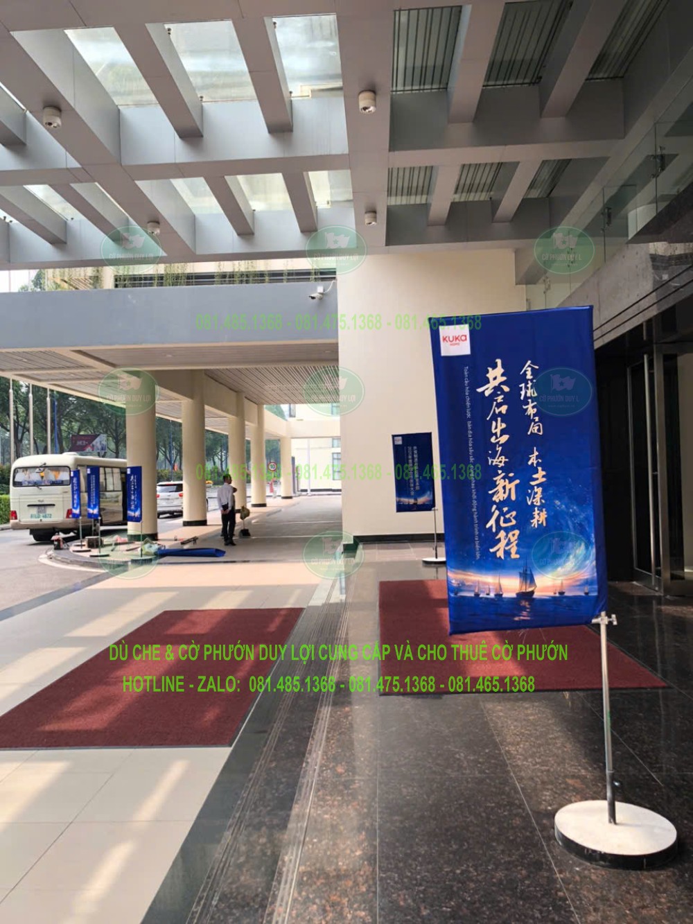 Cờ chữ nhật nền xanh in chữ được dựng bằng đế tròn tại khu vực sảnh có mái che.