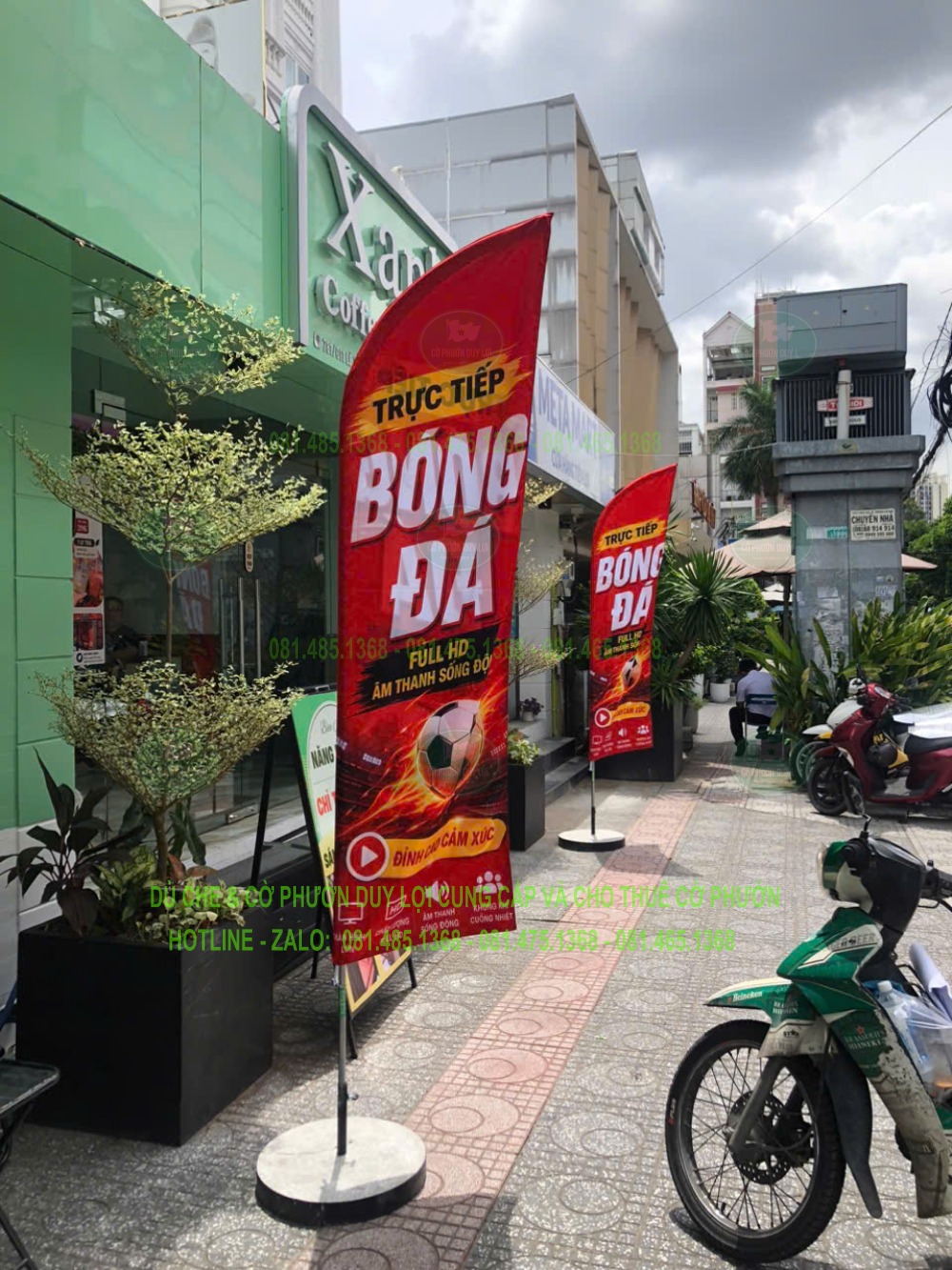 Cờ đỏ có nội dung “Trực tiếp bóng đá” dựng trước mặt tiền quán có bảng hiệu màu xanh.