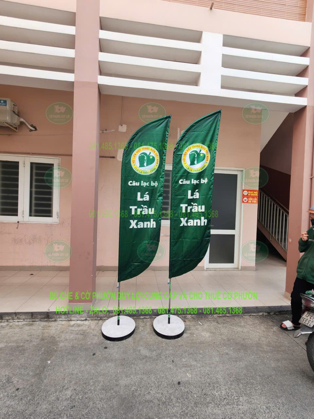 Hai cờ nền xanh mang chữ Lá Trầu Xanh, dựng bằng đế tròn trước khu vực có mái che.