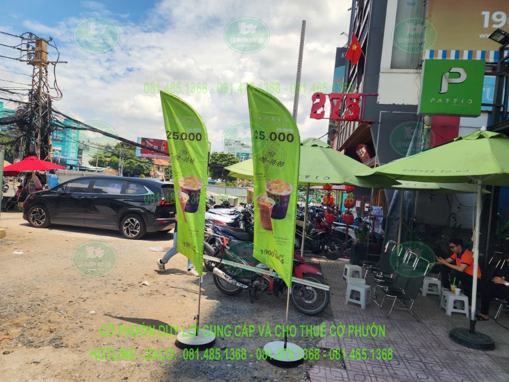Hai cờ nền xanh lá in hình đồ uống, đặt trước quán trong khu vực có bàn ghế và dù che.