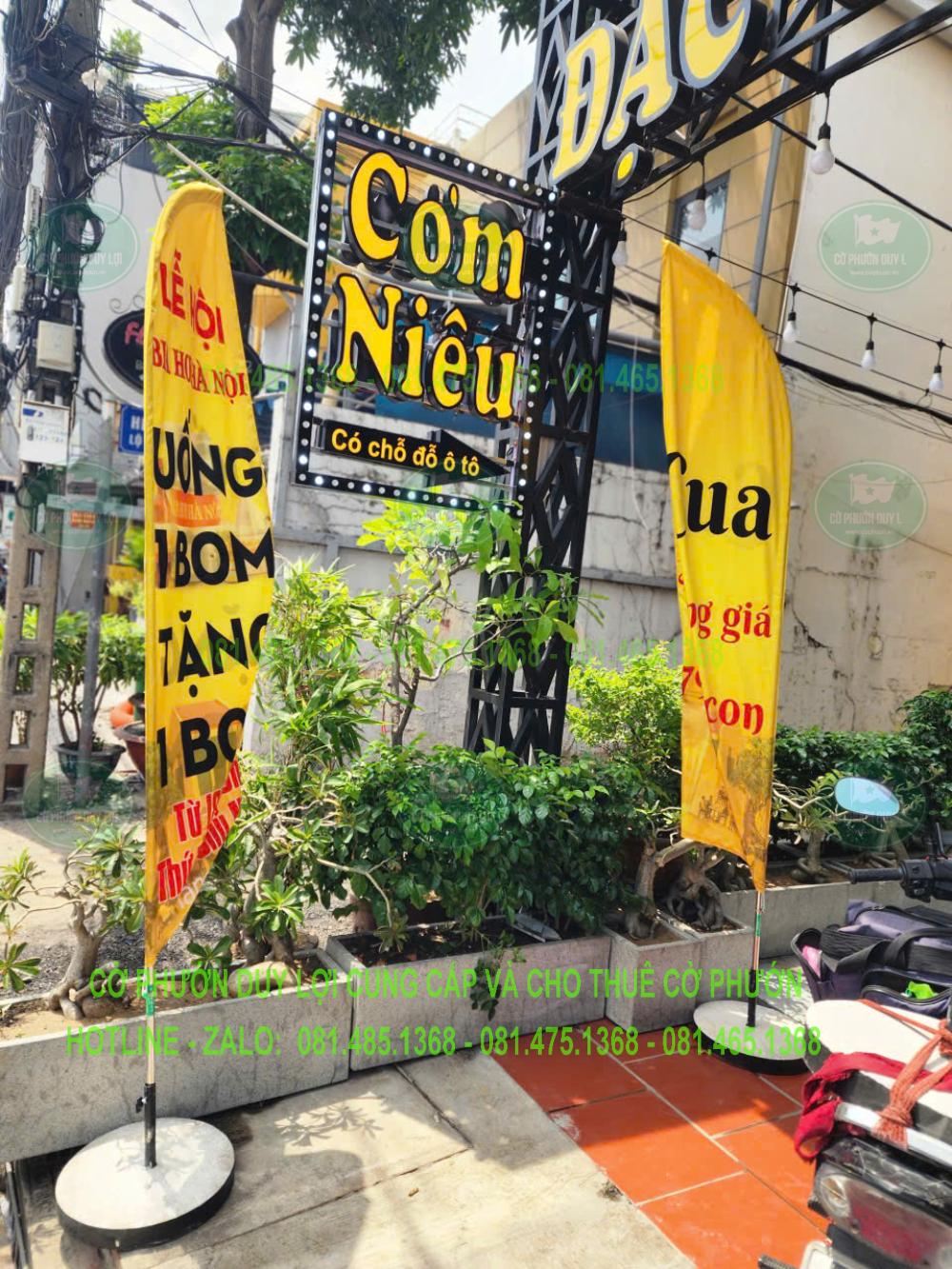 Cờ vàng bố trí gần bảng hiệu Cơm Niêu, với một phần nội dung và đế ở khu vực có cây xanh.